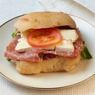 Smörgås i bröd fylld med skivad salami, bitar ost och en tomatskiva på en tallrik.