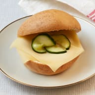 En rund bulle fylld med en ostskiva och gurkskivor på en tallrik.