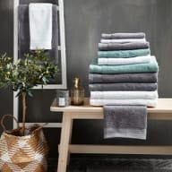 Handdukar ligger staplade på en träbänk bredvid en krukväxt i en flätad korg.