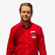 En anställd i butiksklädsel med namnskylt och "Maxi"-logotyp som står framför kameran.