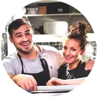 En person i förkläde och en person med uppsatt hår ler i ett kök med hyllor och köksredskap.