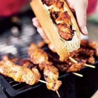 En person håller en baguette fylld med grillat kött ovanför en grillplatta med fler köttspett.