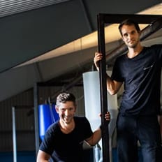 En person står i en rektangulär ram medan en annan person sitter på huk bredvid ramen i vad som ser ut att vara en inomhuslokal med en vattenyta i förgrunden.