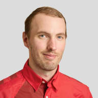 En person med kort hår och skäggstubb bär en skjorta med krage och knappar samt ett namnbricka.