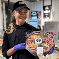 Butiksanställd håller upp en plastförpackad serveringsbricka med räkor och tillbehör framför fiskdisken.