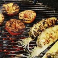 Grillad mat på ett grillgaller över glödande kol, inklusive majskolvar och frukthalvor.