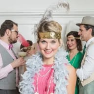 En grupp personer i festkläder från 1920-talet med fjäderboor, glittrande huvudband och hattar står tillsammans inomhus.