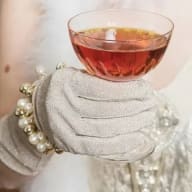 En hand i vante håller ett glas med dryck bredvid ett pärlhalsband.