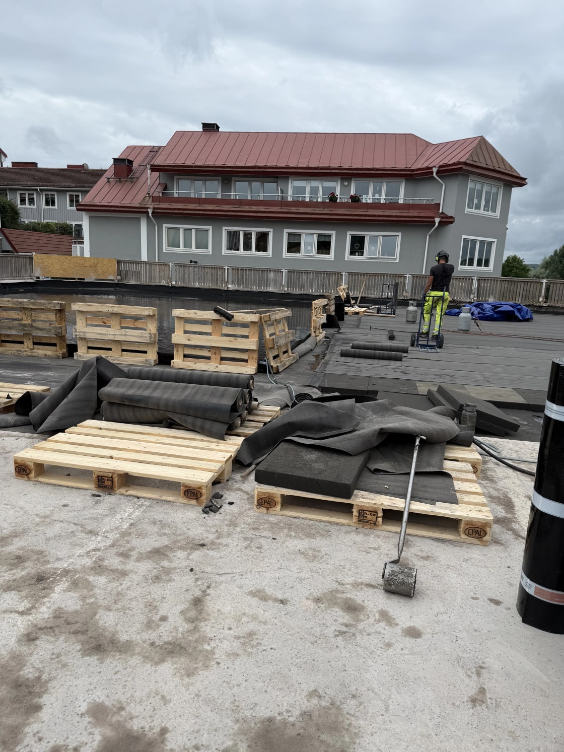 Person arbetar på ett platt tak med takrullar och byggmaterial på träpallar.
