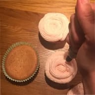 En person dekorerar muffins med frosting medan en ofylld muffins ligger bredvid på ett träbord.
