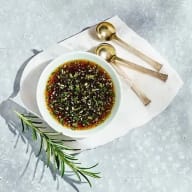 En skål med kryddad olja och örter står på en tallrik med två skedar bredvid och en kvist rosmarin.