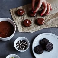 En person lägger chokladöverdragna kakor på ett papper bredvid skålar med chokladsås, strössel och mörka runda kakor.