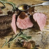 En tallrik med skivor av kött, en sked med sås och kvistar av rosmarin på en träyta.