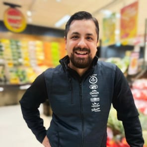 Person i en matbutik står med ena handen i sidan framför frukt- och grönsaksdisk.