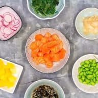 Flera skålar med olika ingredienser står på ett bord, inklusive laxbitar, grönsaker och bönor.