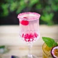 Ett glas med dryck och en körsbär står på ett bord bredvid ett annat glas med en citrusskiva och ett blad.