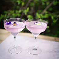 Två cocktailglas med dryck och blommor som garnering står på ett bord utomhus med grönska i bakgrunden.