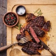 En grillad köttbit ligger på en skärbräda med en gaffel och kniv bredvid, samt två små skålar med kryddor och örter.