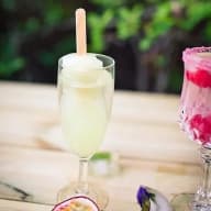 En glasflaska med dryck och pinne står bredvid ett glas med annan dryck på ett bord utomhus.