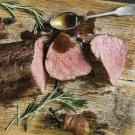 En tallrik med skivor av kött, en sked med sås och kvistar av rosmarin på en träyta.