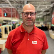 En person med glasögon och skjorta med Maxi-logotyp står i en butik med hyllor i bakgrunden.