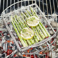 En grillkorg med sparris och citronskivor ligger på ett grillgaller över glödande kol.