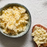 En skål med smulad ost bredvid en brödskiva med pålägg på en bordsyta.