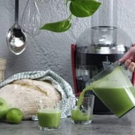 En person häller grön dryck från en kanna till ett glas bredvid en juicepress, bröd och limefrukter på en bänkskiva.