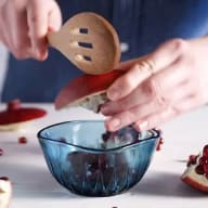 En person håller en sked över en glasskål medan en granatäpple skärs bredvid.