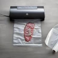 En vakuumförpackningsmaskin står på ett bord bredvid en förseglad plastpåse med ett stycke kött inuti.