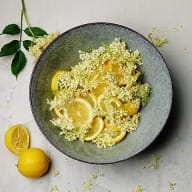 En skål med citronskivor och blommor står bredvid en hel citron på ett bord.