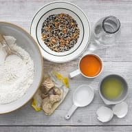En bild visar ingredienser för bakning, inklusive en skål med mjöl, en tallrik med fröblandning, en kopp med vätska, ägg, jäst och en behållare med olja.