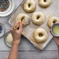En person förbereder bagels på en bakplåt bredvid skålar med olika fröer och en kopp med vätska.