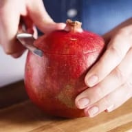 En person skär i ett granatäpple på en träyta med en kniv.