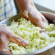 En person blandar strimlad kål i en metallskål med händerna.