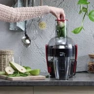 En person häller gröna blad i en juicemaskin bredvid en skärbräda med grönsaker på en köksbänk.