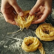 En person formar spaghetti till små nystan på en mörk yta med mjöl.