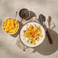 En skål med yoghurt och fruktbitar står på ett bord tillsammans med en skål mango, en behållare med frön och en sked.