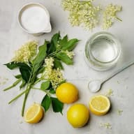 En bild visar citroner, gröna blad, små vita blommor, en skål med vätska och ett glas med vätska på en ljus yta.