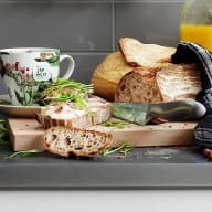 En frukostscen med bröd, en smörgås, en mugg och gröna växter på en skärbräda.