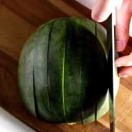 En person skär en vattenmelon med en kniv på ett träbord.
