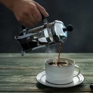 En person häller kaffe från en kanna till en kopp på ett träbord.