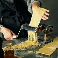 En person använder en pastamaskin för att göra färsk pasta på ett bord med en deg och en flaska olja bredvid.