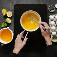 En person vispar en gul blandning i en skål bredvid citrusfrukter, ägg och en kopp med vätska.