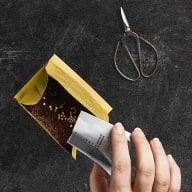 En person håller en kniv mot en öppnad förpackning bredvid en sax på en mörk yta.