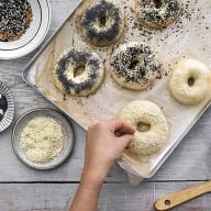 En person strör frön på en bagel på en bakplåt med flera andra bagels, bredvid en skål med frön och en träsked.
