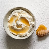 En skål med krämig massa och honung, bredvid en honungssked på en vit yta.