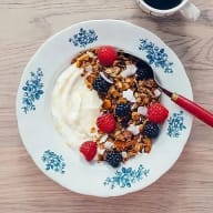 Frukosttallrik med yoghurt, granola och färska hallon och blåbär, sked och en kopp kaffe.