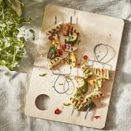 En skärbräda med grillad zucchini och andra grönsaker ligger på ett bord med en duk och några örter bredvid.