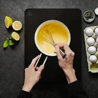 En person vispar en gul smet i en skål medan citrusfrukter, ägg och kryddor ligger bredvid på en mörk yta.